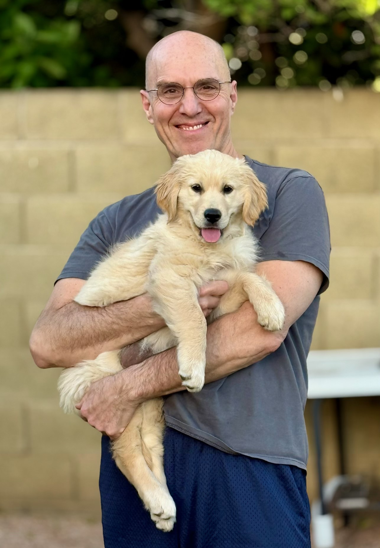 A man wearing glasses, a gray t-shirt, and blue shorts stands outside holding a light-colored puppy in his arms. The man is smiling, and the puppy has its tongue out. A tan brick wall is in the background.