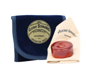 A round amber-colored Bernadel Colophon sits on a beige cloth beside a dark blue velvet pouch, both labeled "Gustave Bernardel Colophane.
