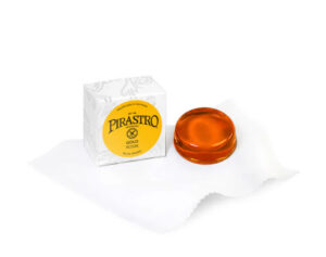 A round, amber Pirastro Goldflex Viola Rosin rests on a white cloth beside its white and yellow box, which displays gold lettering and decorative accents.